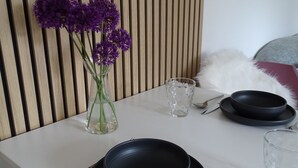Una mesa de comedor con platos negros, cubiertos y una copa de vidrio transparente. Un jarrón con flores moradas está sobre la mesa.
