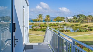 A view from a balcony overlooking a landscaped garden with a pond, palm trees, and a distant body of water.