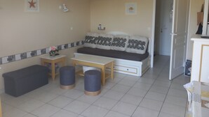 Una sala de estar con un sofá, dos otomanas, una mesa de centro y un banco. Hay dos mesitas con adornos decorativos. El piso es de baldosas y las paredes son color beige.