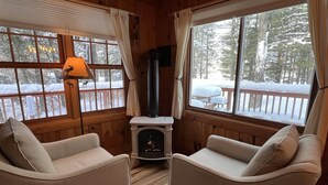 A cozy cabin interior with two beige armchairs, a fireplace, and large windows offering a snowy forest view.