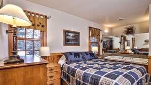 A bedroom with a large bed, a wooden dresser, a desk with a lamp, and a window with a view of snow-covered trees.