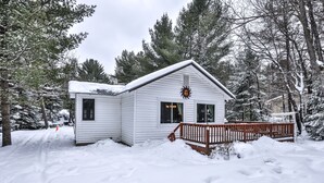A white cabin with a wooden deck, surrounded by snow-covered trees.