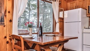 A wooden cabin interior with a dining table set for two, a white refrigerator, and a window with a view of trees.