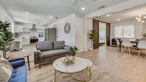 A spacious living area with a sofa, coffee table, and dining table. There is a kitchen in the background with cabinets and a stove.