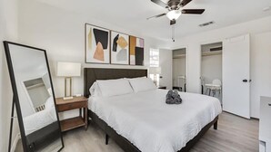 A modern bedroom with a large bed, a ceiling fan, a bedside table with a lamp, and abstract wall art.
