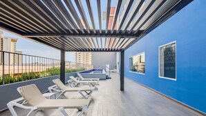 A covered outdoor lounge area with sun loungers, a pool, and a view of the cityscape. - One Mazatlán (Mazatlán)