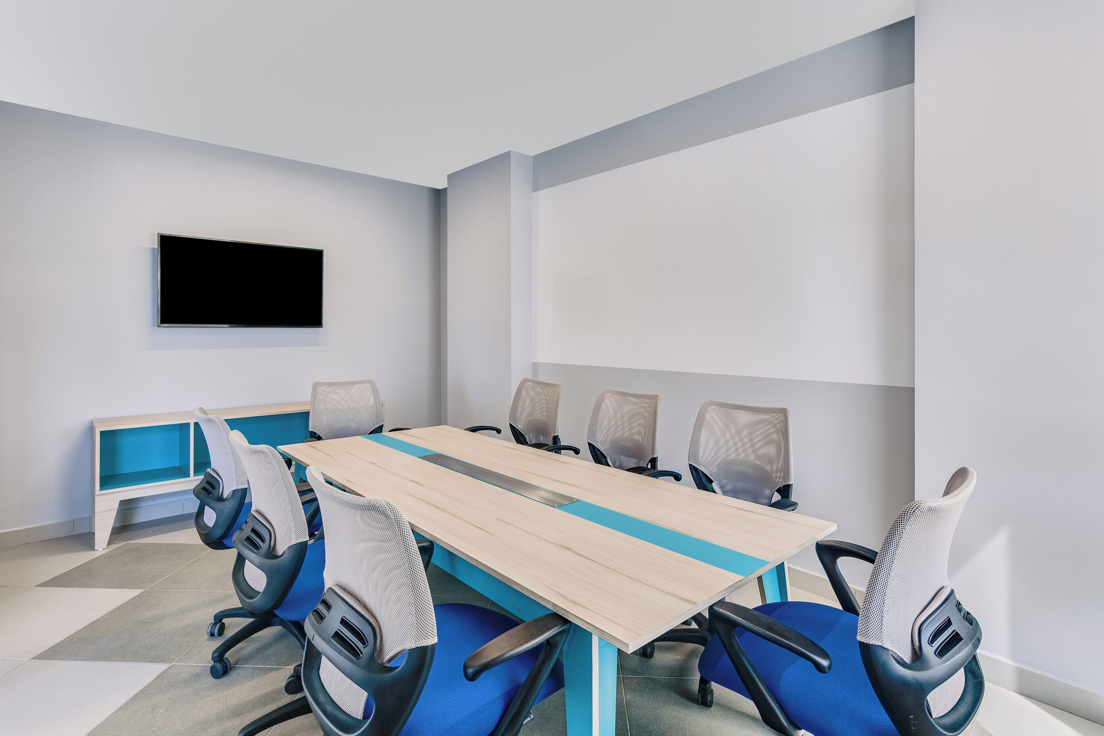 A conference room with a long table, six chairs, a TV mounted on the wall, and a blue bench against the wall.