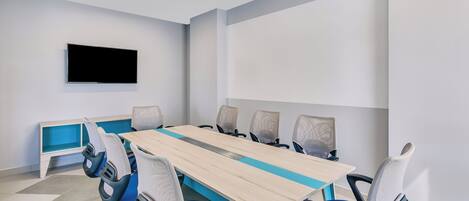 A conference room with a long table, six chairs, a TV mounted on the wall, and a blue bench against the wall.