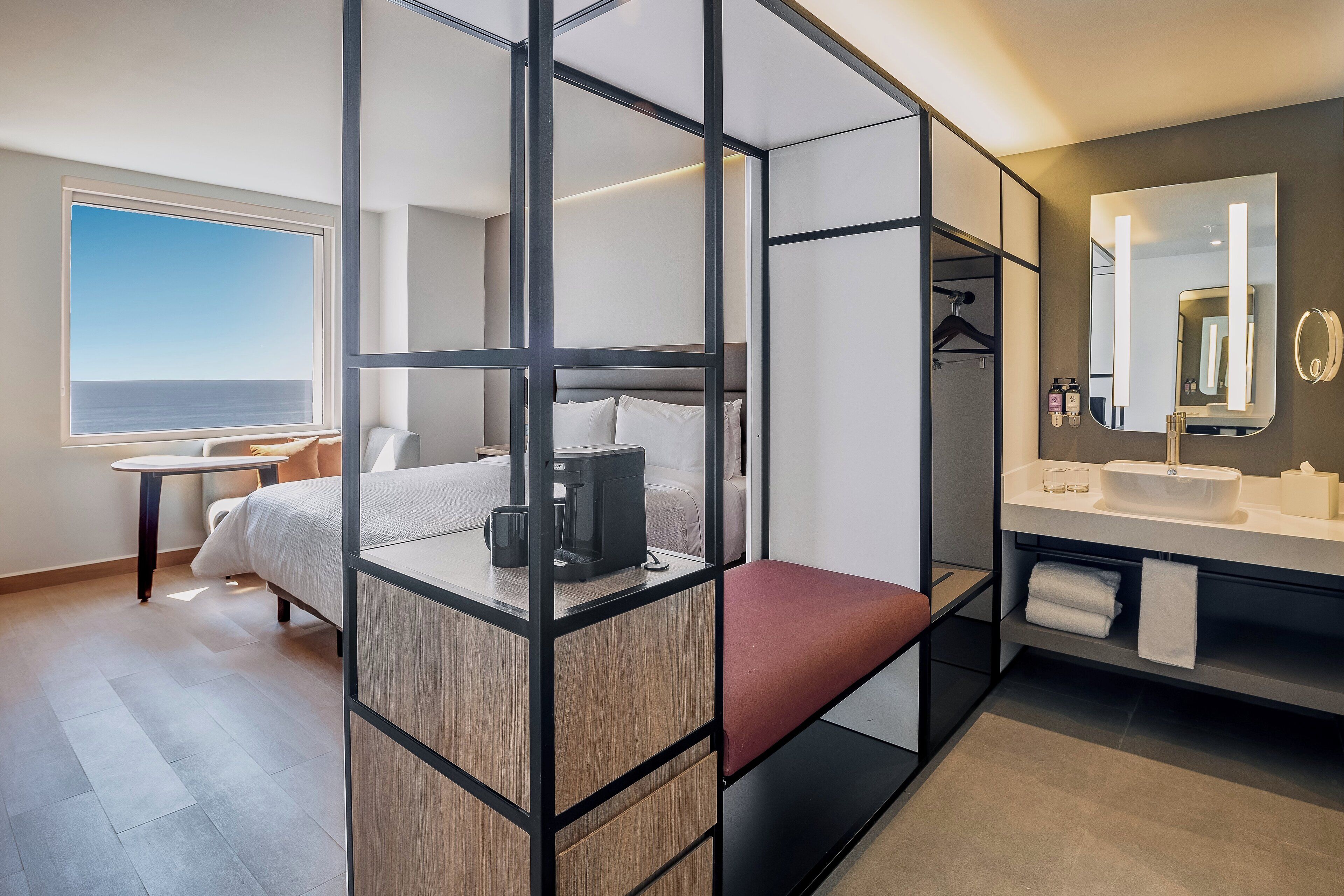 Habitación de hotel moderna con una cama grande, un escritorio, un área de descanso, un baño con espejo y vista al mar.