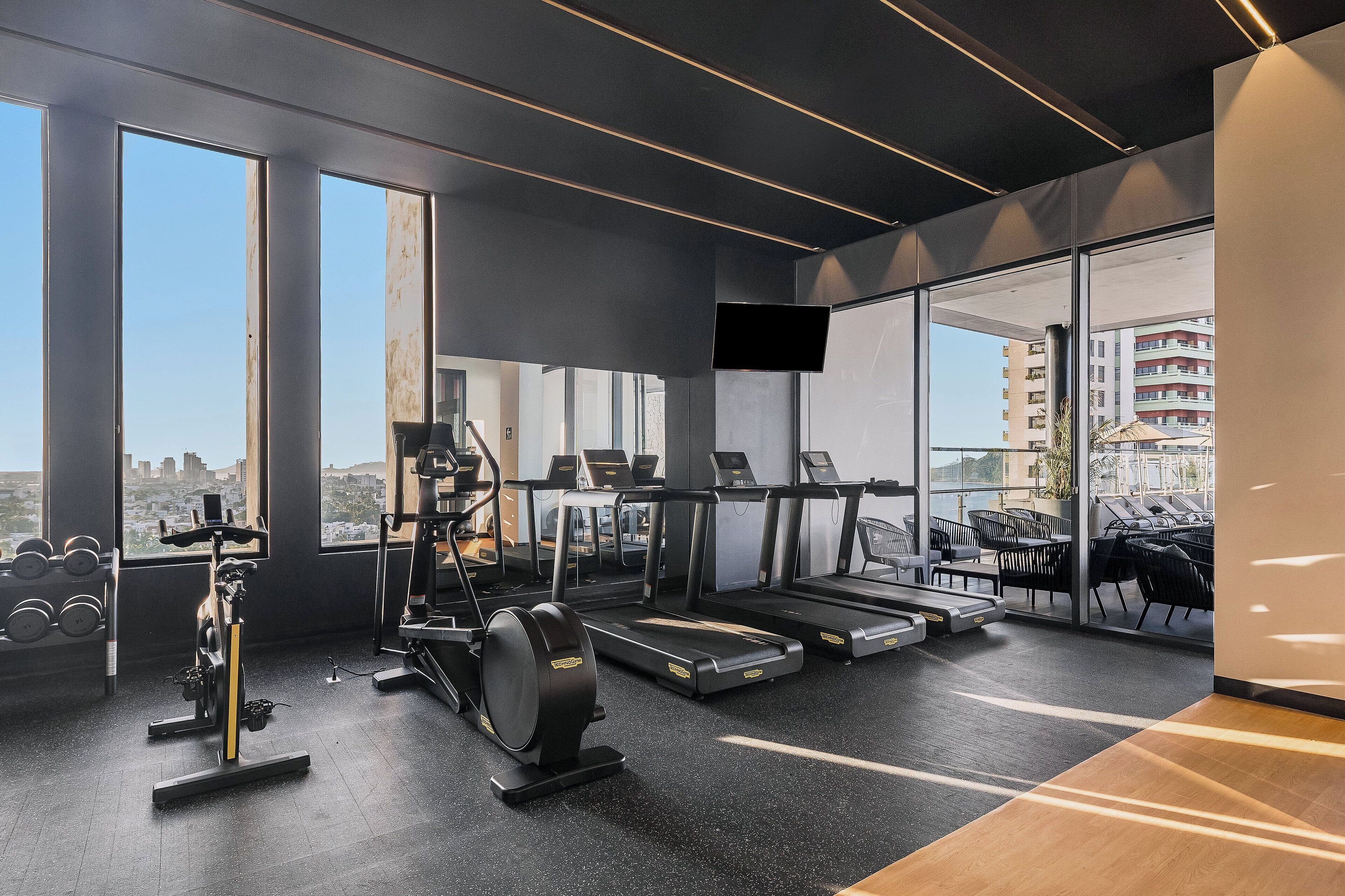A modern gym with treadmills, an elliptical, and dumbbells, large windows, and a view of the city.