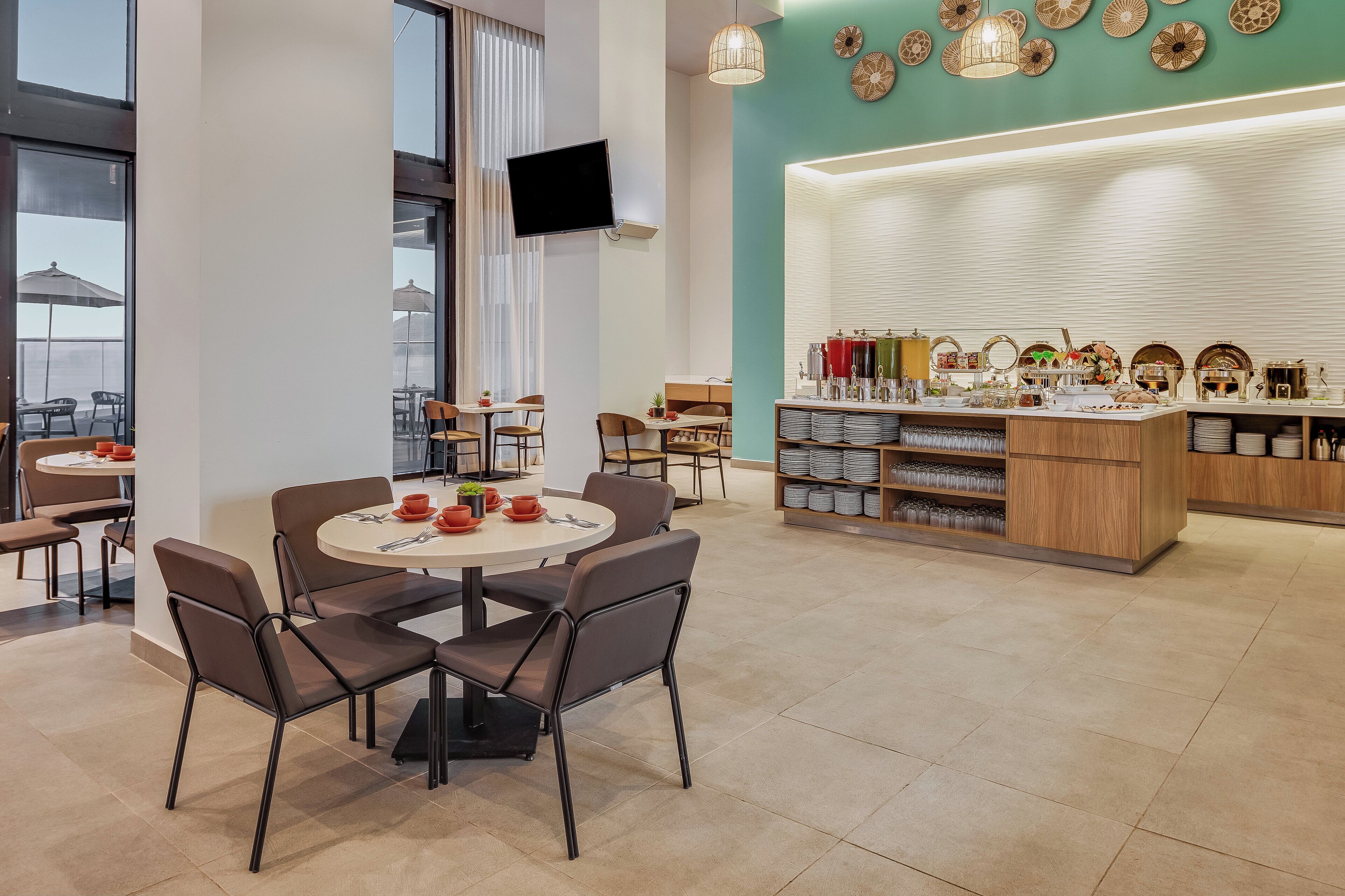 A modern dining area with round tables and chairs, a kitchen counter with various items, and a large window overlooking the city.