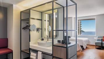 A modern bathroom with a large mirror, a sink, and a shower area. There is a bed and a chair in the adjacent room.