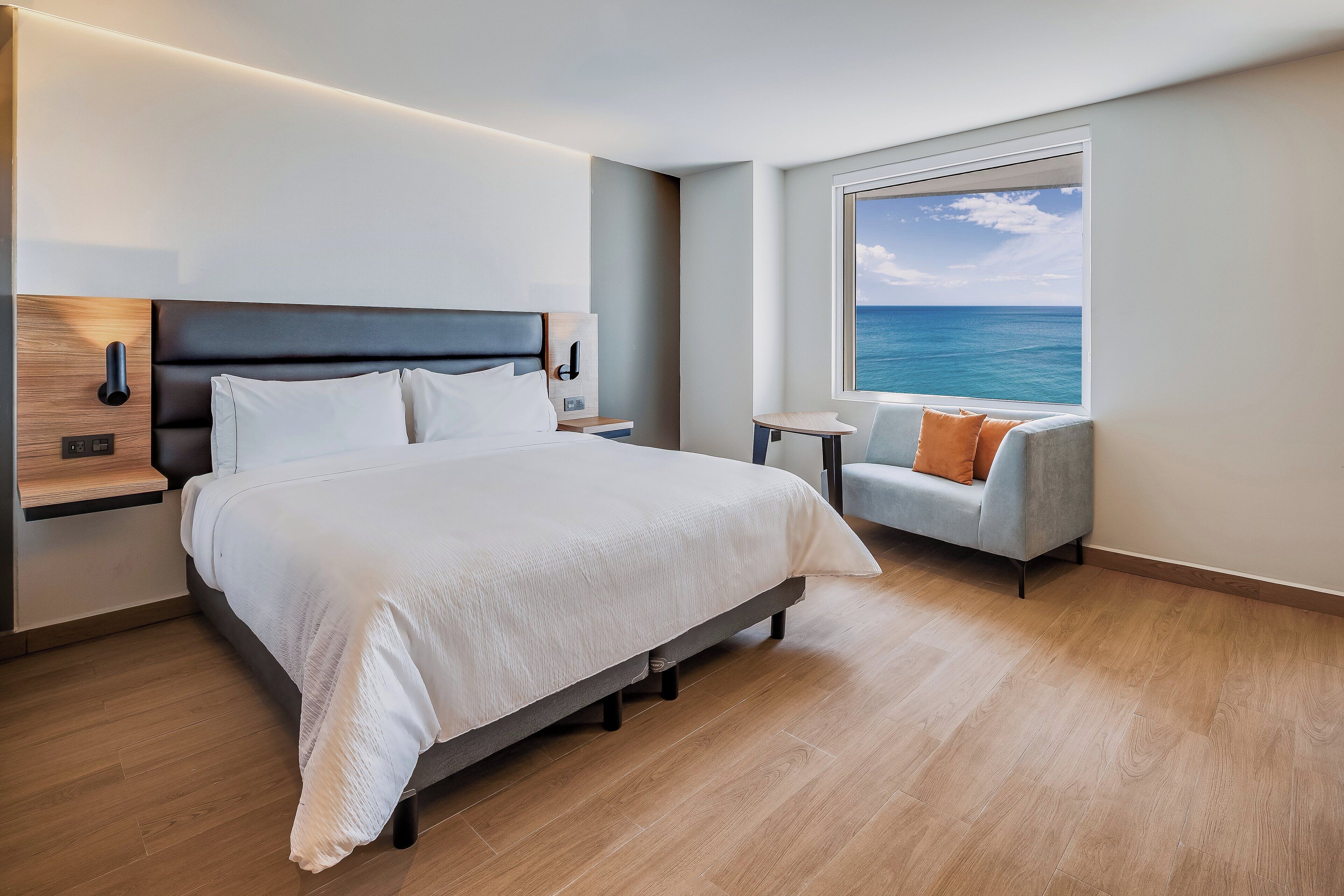 A modern hotel room with a large bed, a small table, a sofa, and a window with a view of the sea.