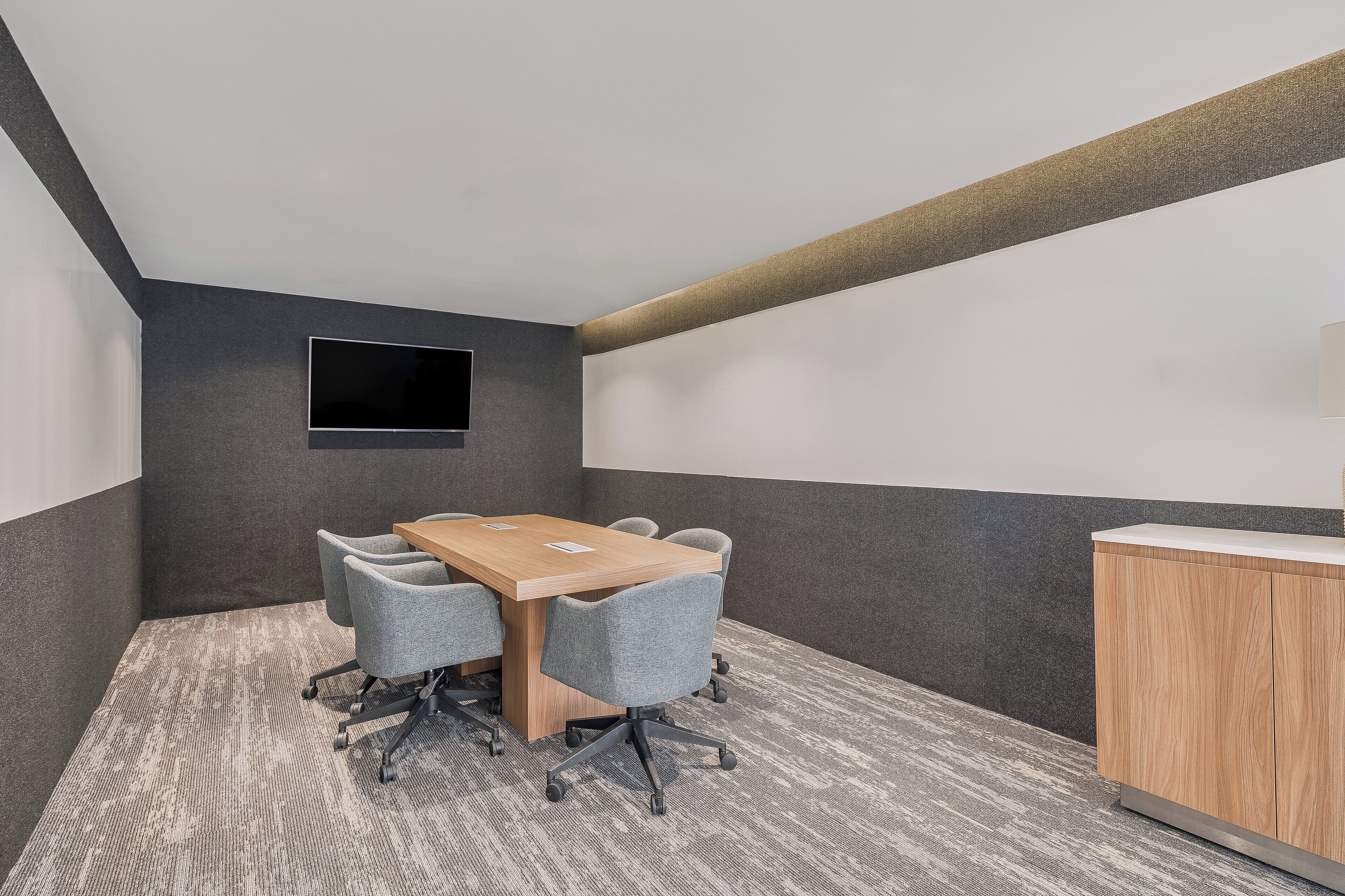 Un salón de conferencias con una mesa de madera, sillas grises, un televisor pequeño en la pared y un armario de madera.