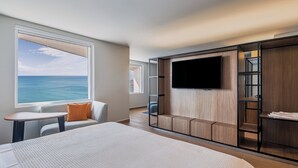 Une chambre d’hôtel moderne avec un grand lit, un coin salon offrant une vue sur l’océan, un téléviseur fixé à un mur en bois et une petite table.