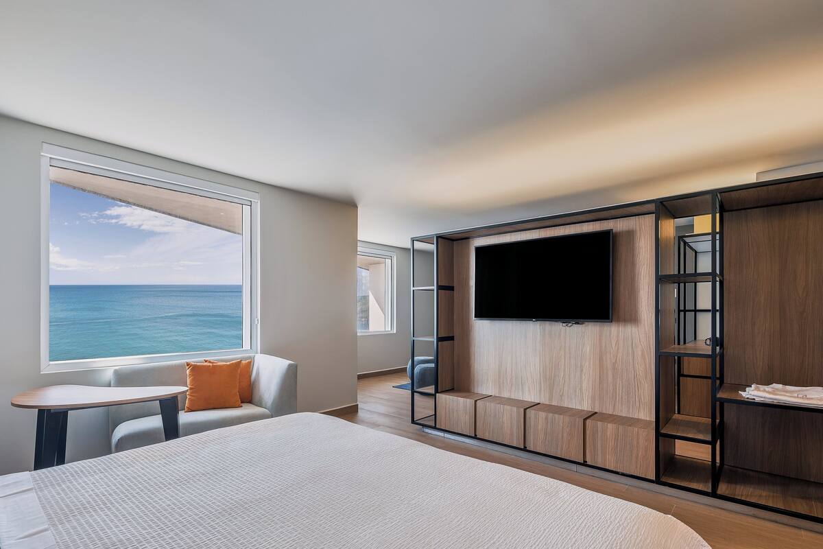 A modern hotel room with a large bed, a seating area with a view of the ocean, a TV mounted on a wooden wall, and a small table.