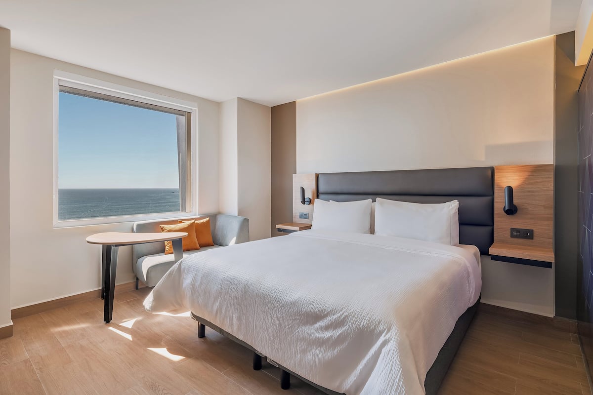 A modern hotel room with a large bed, a small table, a chair, a nightstand, and a view of the sea.