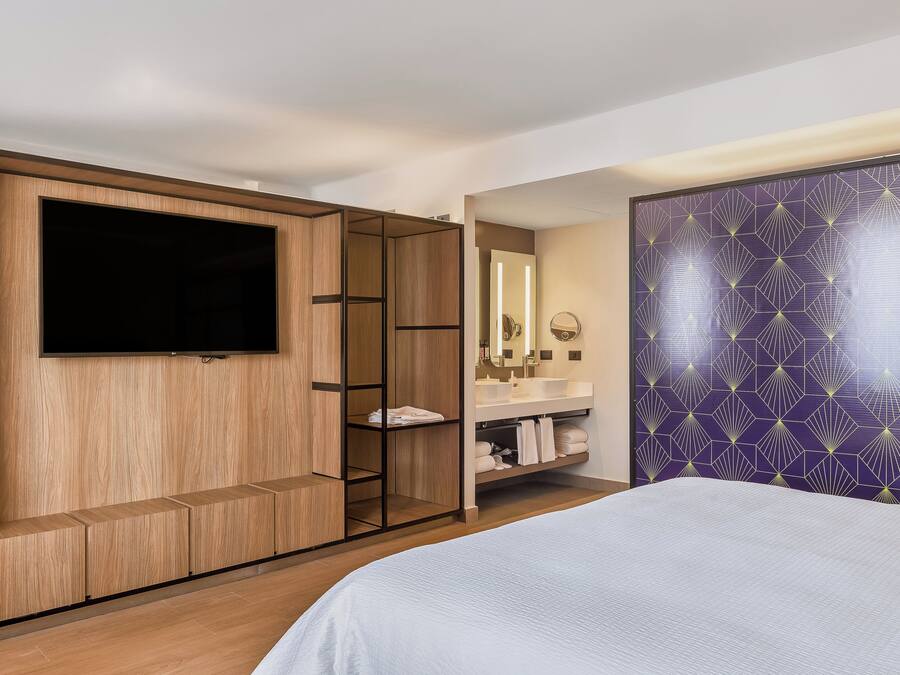 Quarto de hotel com uma cama grande, uma TV fixada num armário de madeira, um banheiro com espelho e pia, e uma parede com padrão roxo.
