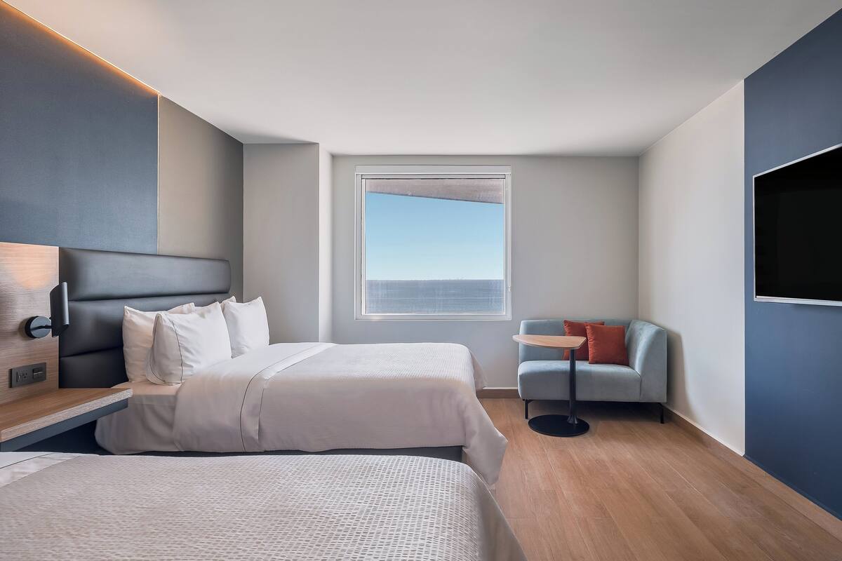 A modern hotel room with two beds, a small seating area, a TV, and a window with a view.