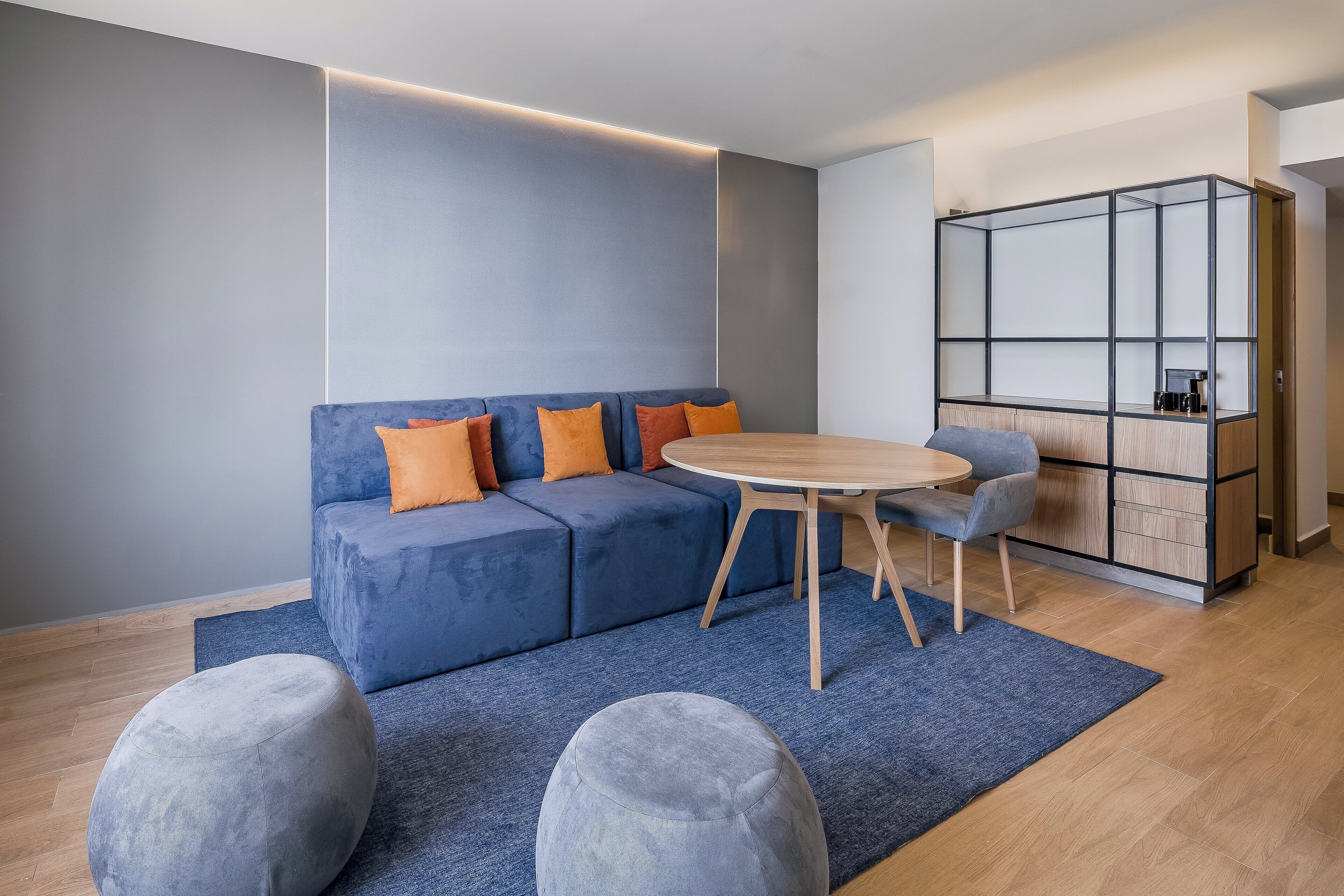 A modern living room with a blue sofa, orange cushions, a round wooden table, and a grey ottoman.