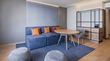 A modern living room with a blue sofa, orange cushions, a round wooden table, and a grey ottoman.