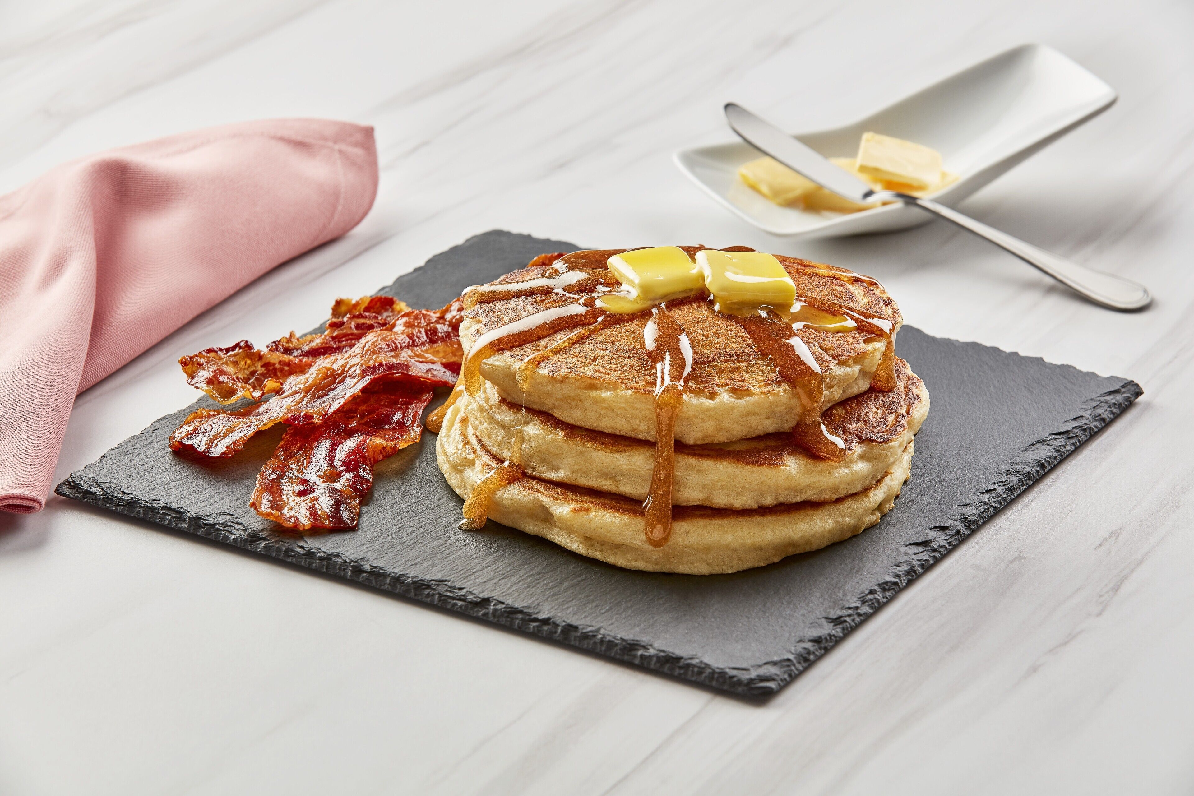 Una pila de tortitas con almíbar y mantequilla sobre un plato de pizarra, acompañada de tiras de tocino y un pañuelo rosa.