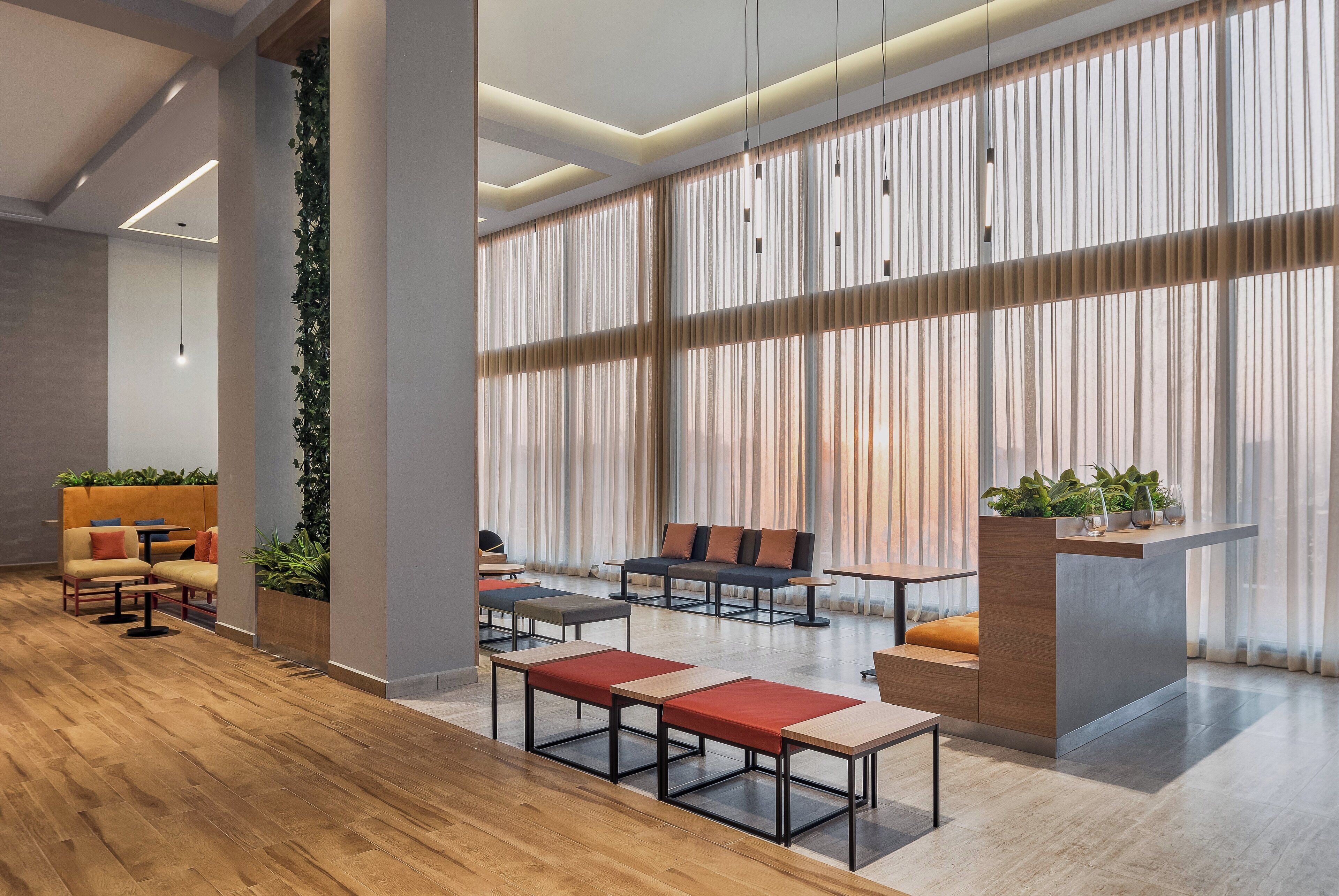 A modern lounge area with wooden flooring, seating arrangements, and large windows with sheer curtains.