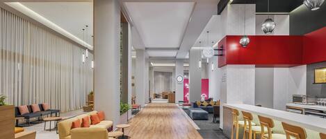 A modern hotel lobby with a long wooden floor, seating areas, a bar with stools, and a kitchen area.