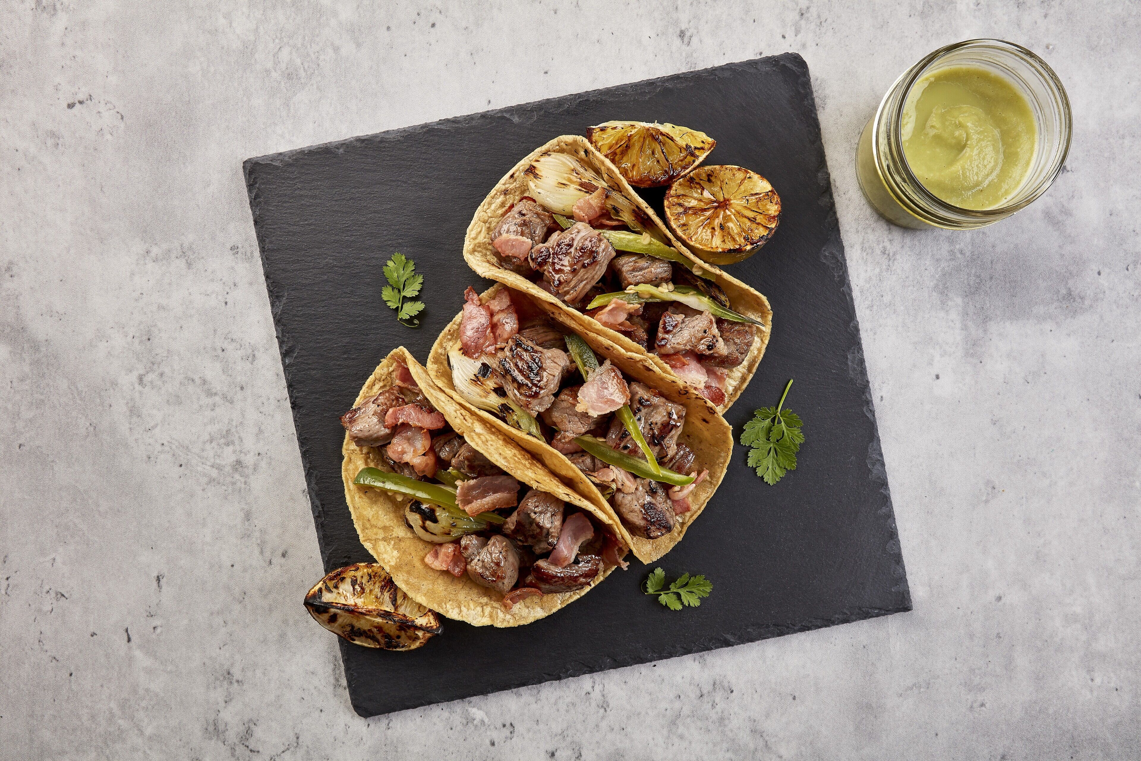 Three tacos with meat and vegetables on a slate board.