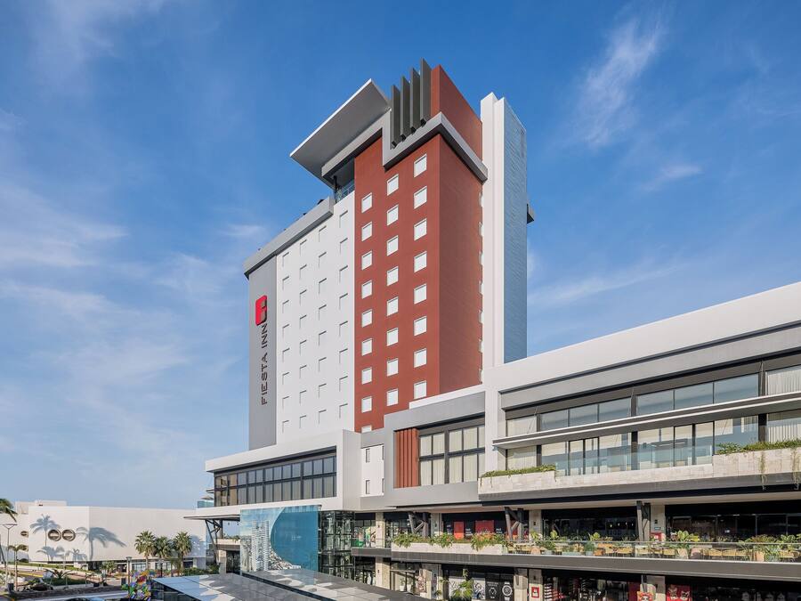 Un hotel moderno con una fachada destacada de rojo y blanco, varios pisos y una terraza en la azotea con asientos al aire libre.