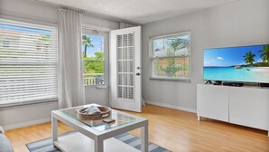 Un salon moderne avec une table basse en verre, un téléviseur posé sur un meuble blanc et de grandes fenêtres équipées de stores.