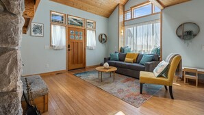 A cozy living room with a wooden ceiling, a stone wall, a sofa with cushions, a coffee table, and a bench. - Seashell Cottage w/ Ocean Views, Hot Tub & Game Room: Ideal for Family & Pets (Blaine)