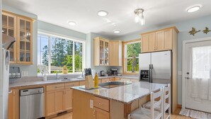 A modern kitchen with wooden cabinets, a stainless steel dishwasher, a large island with bar stools, and a view of the outdoors. - Seashell Cottage w/ Ocean Views, Hot Tub & Game Room: Ideal for Family & Pets (Blaine)