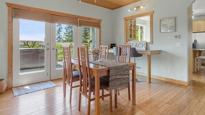 A dining area with a wooden table, chairs, and a chandelier. There is a sliding glass door leading to a balcony. - Seashell Cottage w/ Ocean Views, Hot Tub & Game Room: Ideal for Family & Pets (Blaine)