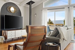 A modern living room with a large flat-screen TV, a wooden chair, a sofa, and a view of the outdoors through large windows. - Stylish & Cozy Lakeview Haven Near Lake, Trails & Mt. Baker Skiing (Bellingham)