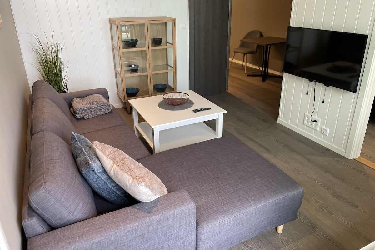 Un salon moderne avec un canapé d’angle gris, une table basse blanche, un meuble vitré contenant des bols, un fauteuil gris et un téléviseur à écran plat fixé à un mur blanc.