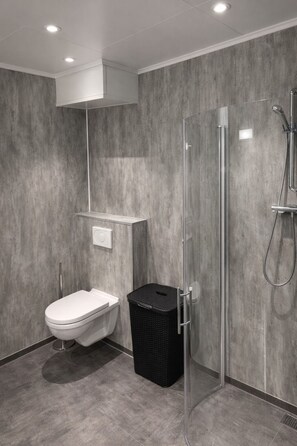 A modern bathroom with a toilet, a black trash bin, a glass shower enclosure, and a white sink. - Arctic Sea Apartments (Hammerfest)