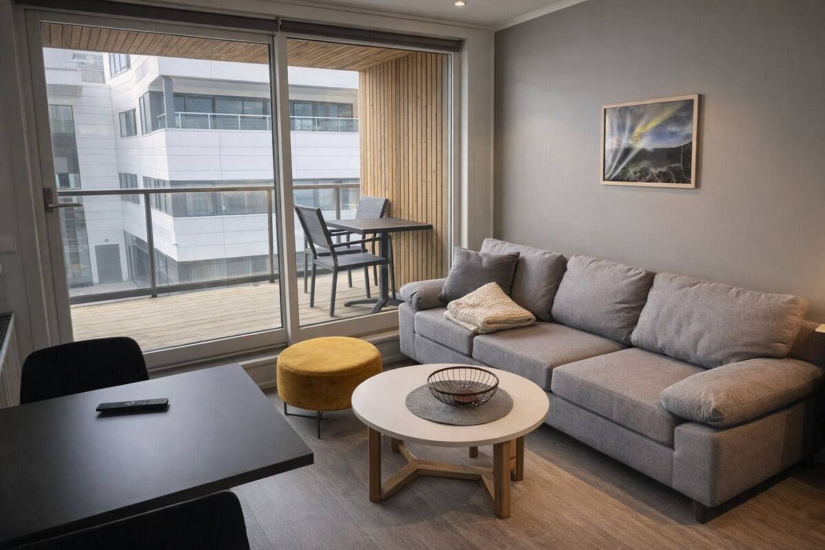 Un salon moderne avec un canapé gris, une petite table ronde et un pouf jaune. Il y a un balcon avec vue sur un immeuble.