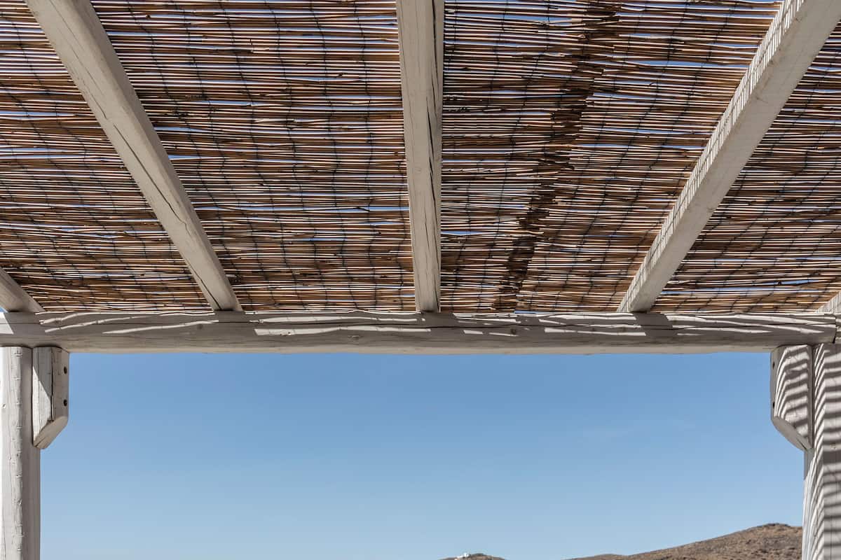 A wooden table and chairs under a pergola with a view of a lake and hills.