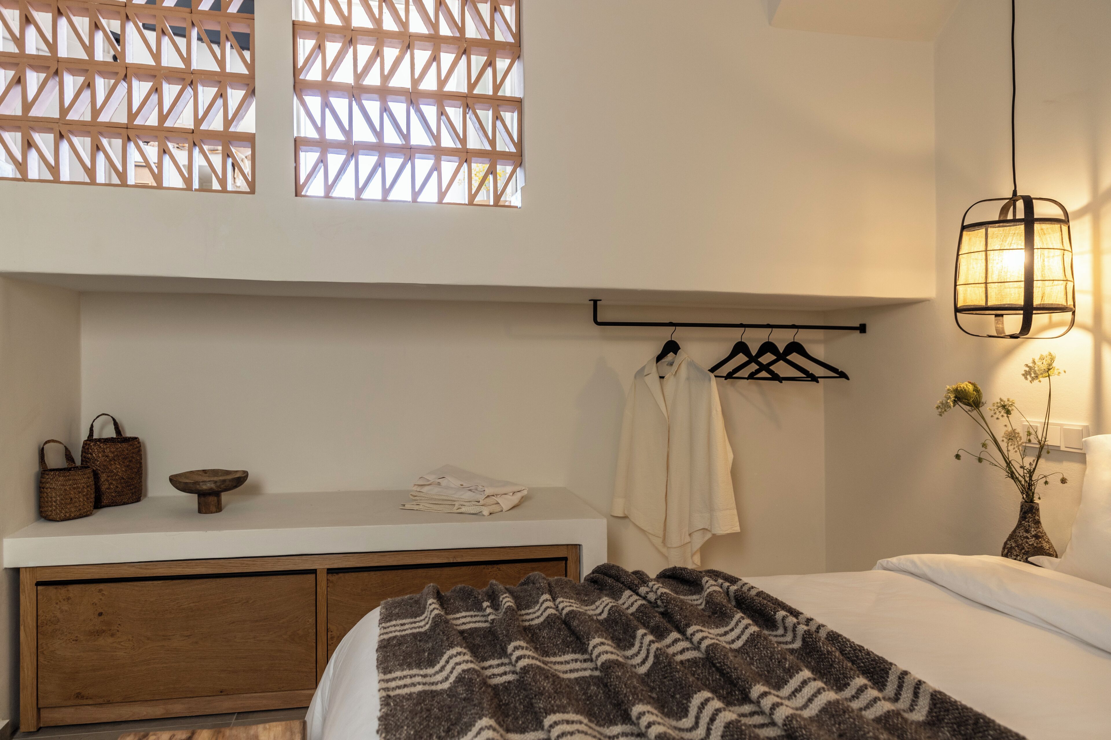A bedroom with a bed, a bedside table, a hanging light fixture, a window with a wooden lattice, and a wall-mounted towel rack.