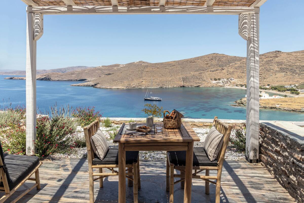 A wooden table with chairs and a basket of bread, overlooking a calm sea and a hilly landscape.