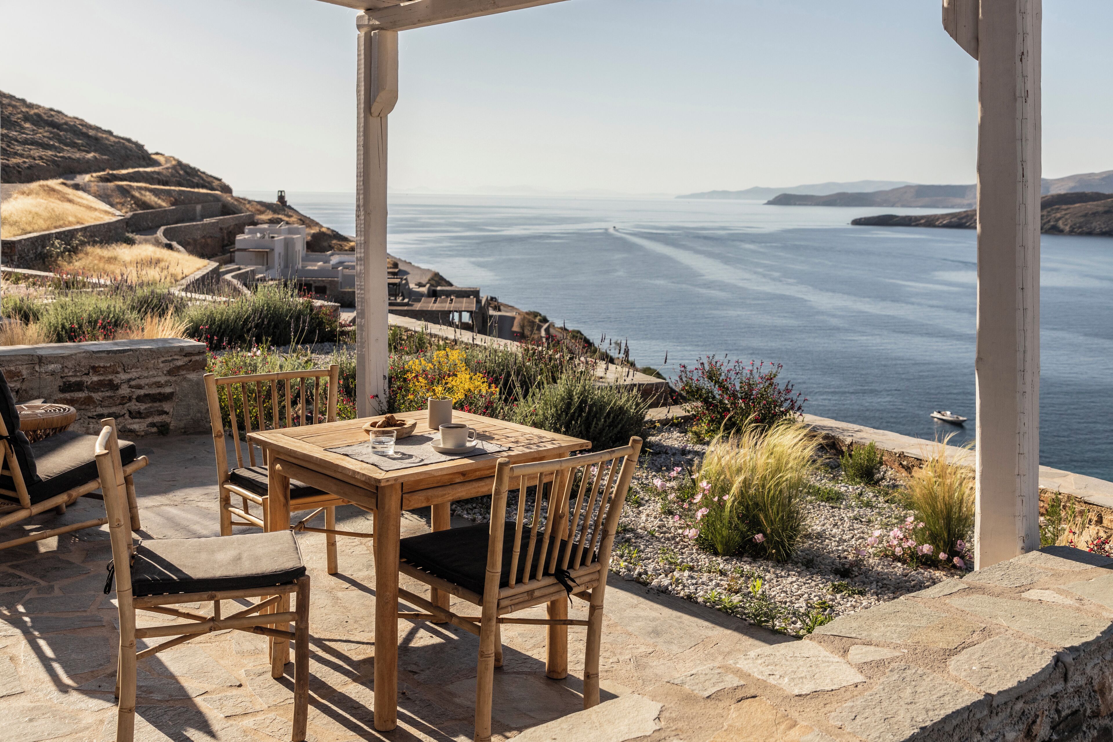 A wooden table and chairs set on a stone patio overlooking a coastal view with a clear sky.