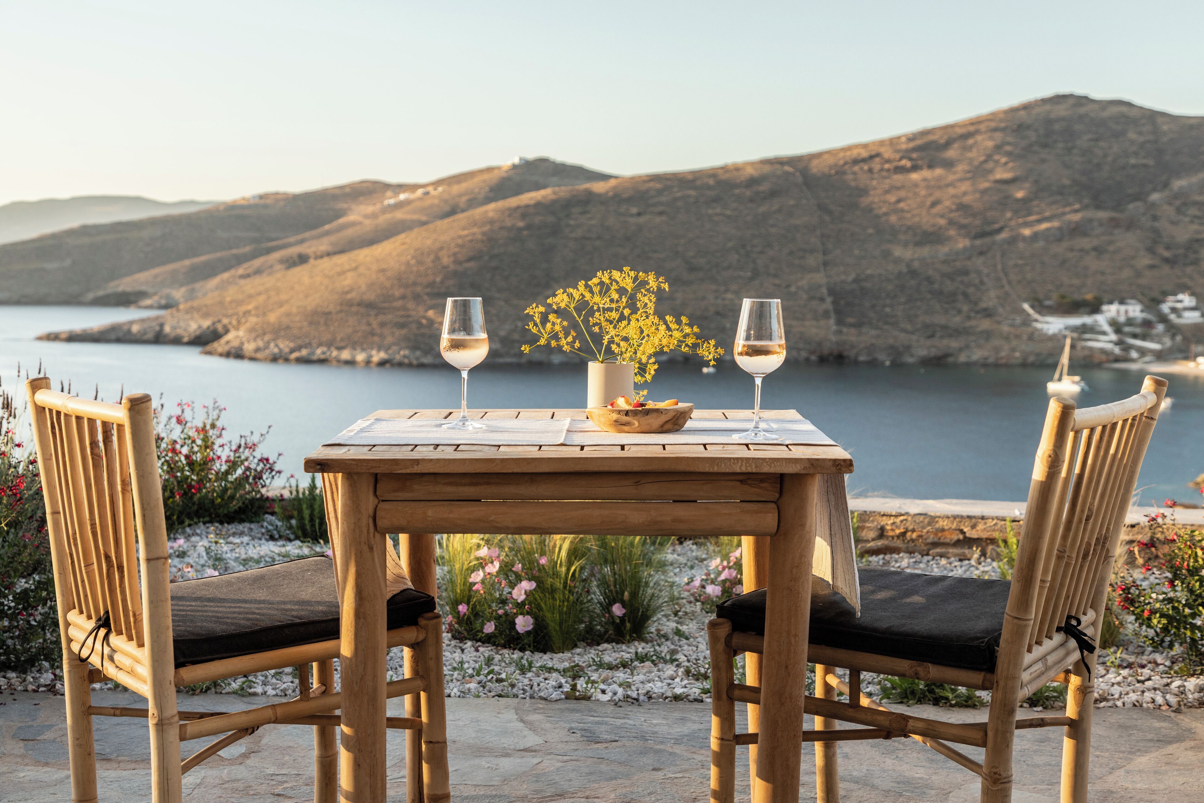 A wooden table with two chairs, a vase with flowers, and two wine glasses set against a scenic backdrop of hills and a body of water.