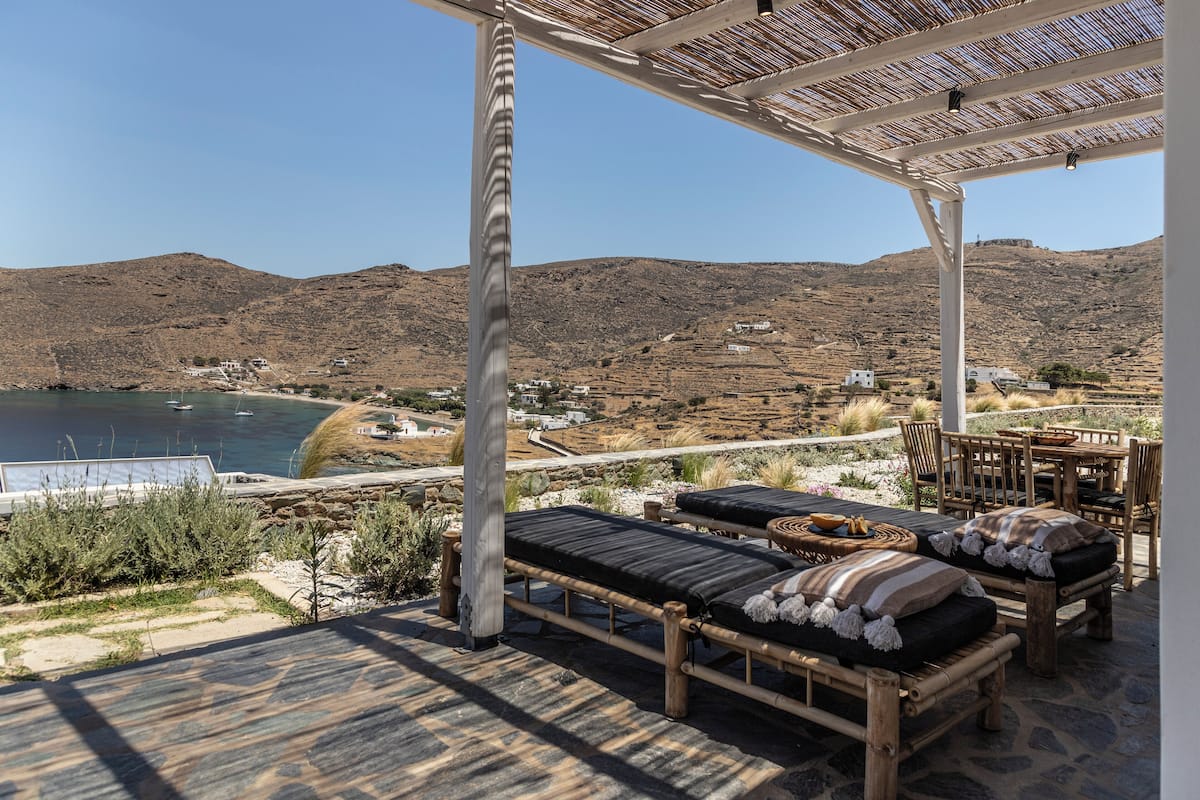 A wooden deck with lounge chairs and a table, overlooking a mountainous landscape and a body of water.