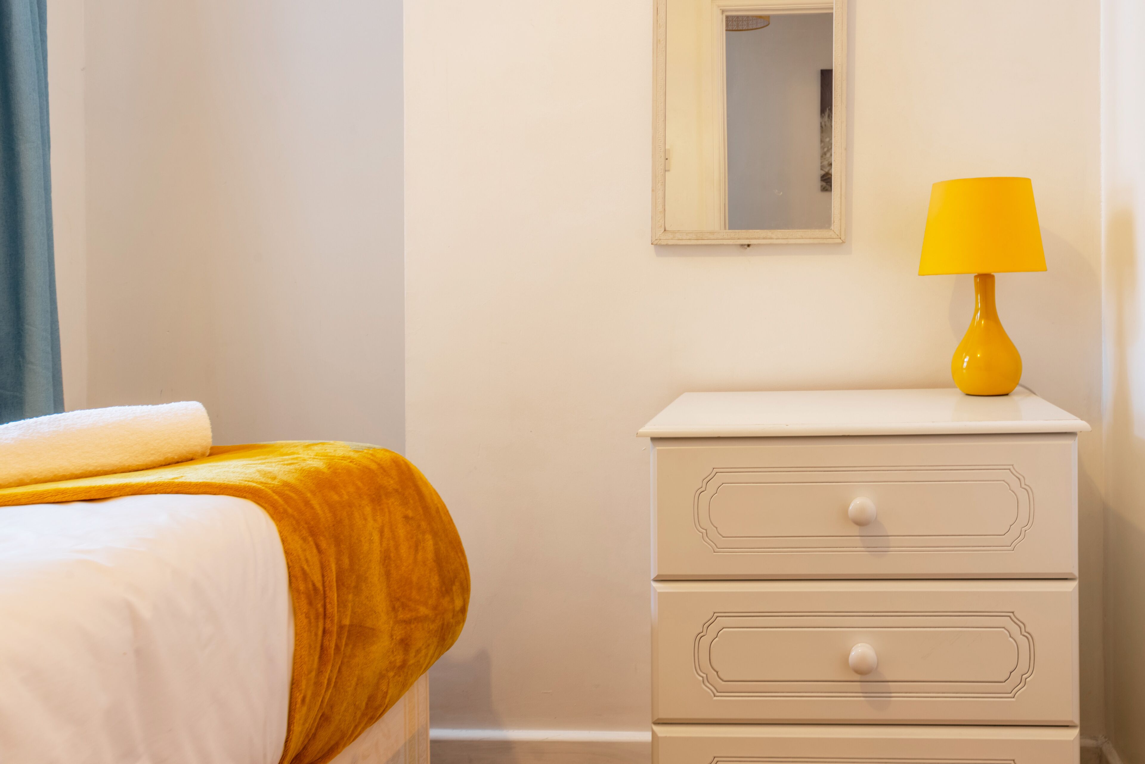 A neatly made bed with a white and yellow blanket, a white bedside table with drawers, and a yellow lamp.