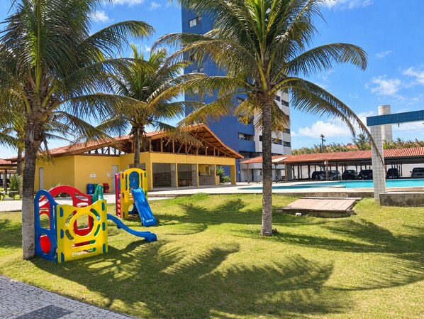 Un'area giochi con scivoli e strutture di arrampicata, una piscina, palme e un edificio con tetto rosso.