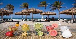 A beach setting with thatched umbrellas, a table with various drinks and fruits, and a view of the ocean and palm trees. - Apartamento 2 suítes lindo e vista mar (Fortaleza)