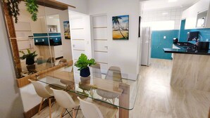 A modern kitchen with a glass dining table, white chairs, and a potted plant. - Apartamento 2 suítes lindo e vista mar (Fortaleza)