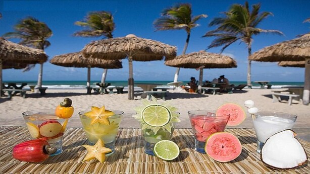 A beach setting with thatched umbrellas, a table with various drinks and fruits, and a view of the ocean and palm trees. - Apartamento 2 suítes lindo e vista mar (Fortaleza)