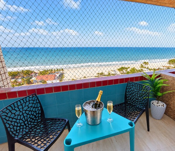 A balcony with a view of the beach, a table with a bottle of champagne, two glasses, and two chairs.
